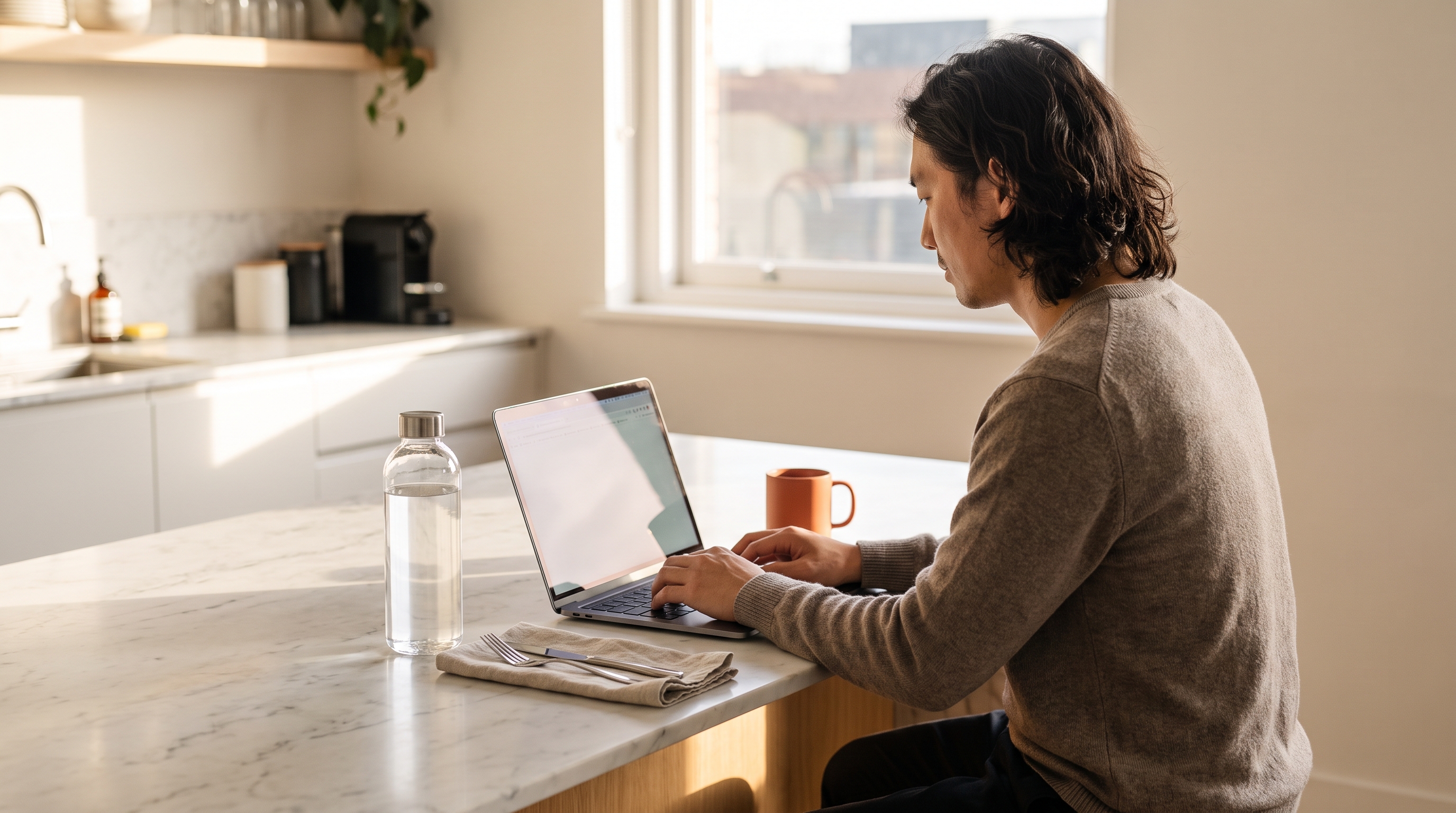 A man in his 30s working focused at a laptop on a kitchen table