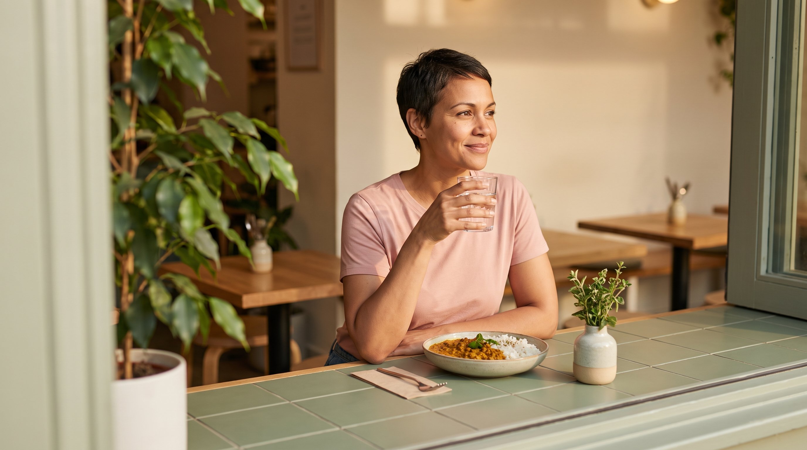 A woman in her 30s relaxing with a glass of water after a meal at a cozy breakfast table