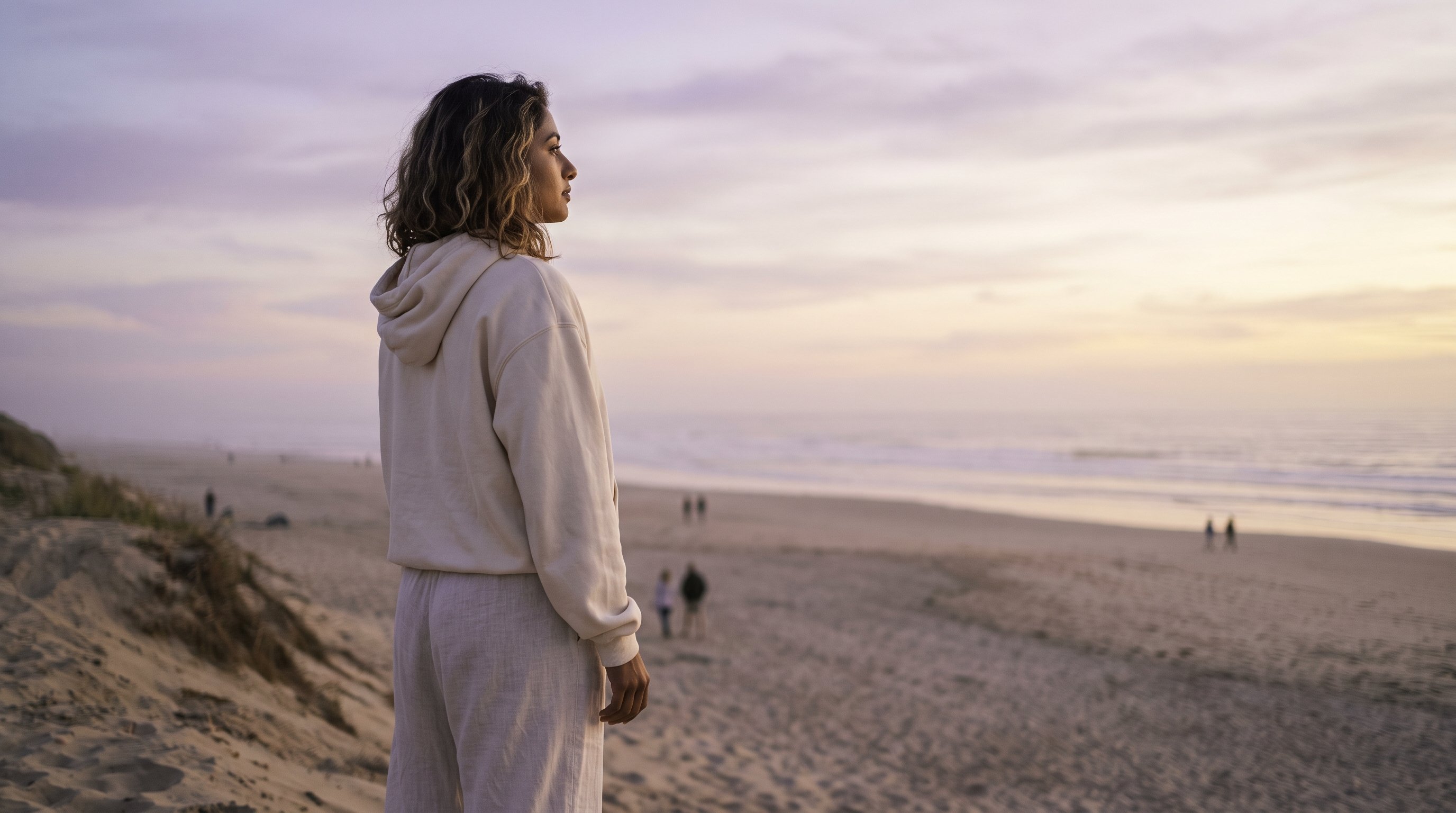 A woman in her late 20s in light flowing clothes gazing at a soft evening sky from a hilltop