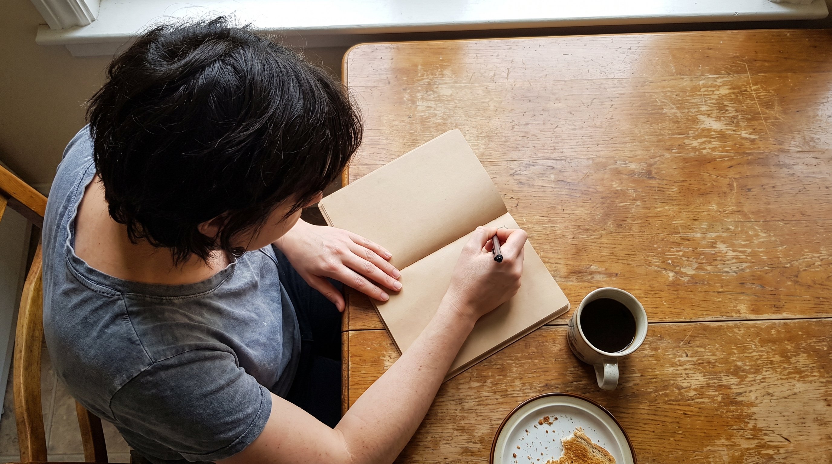 Woman writing in a small notebook at a kitchen table in the morning