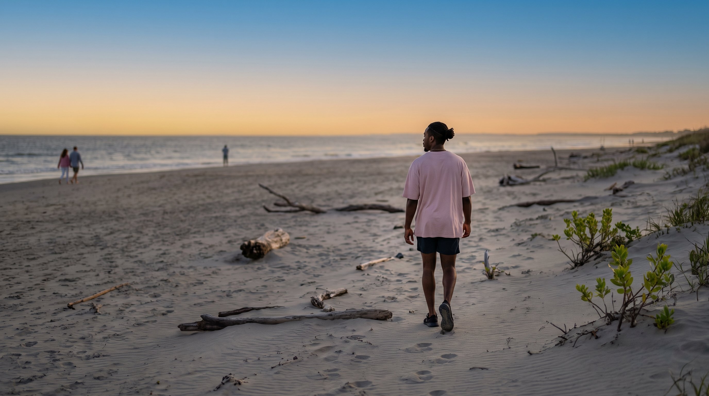 A man in his late 20s walking along a quiet beach at dusk