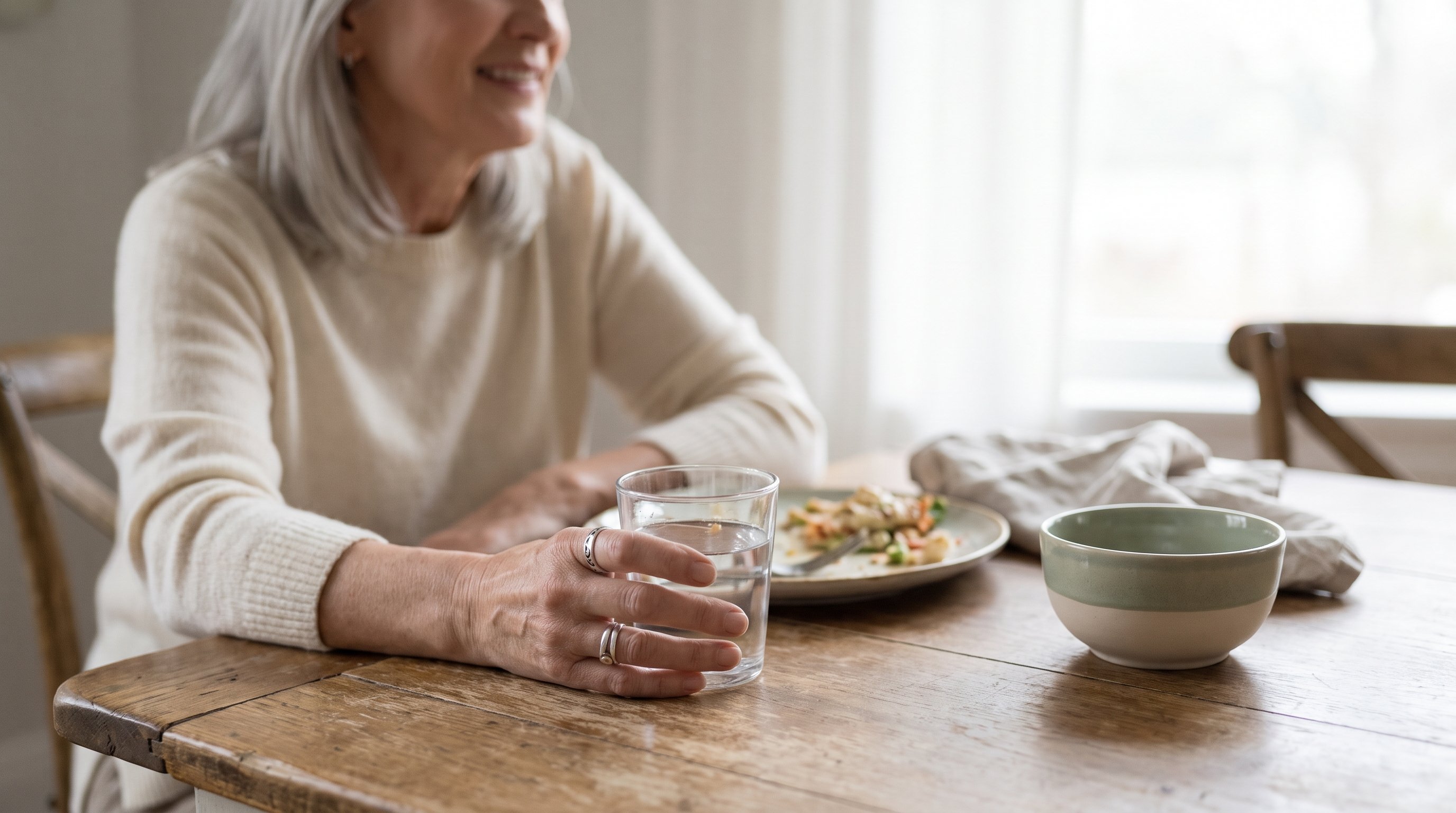 A woman in her 60s with silver hair relaxing with a glass of water after a meal at a calm dining table